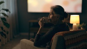 Mulher idosa sentada na poltrona tomando café enquanto assiste televisão numa sala à noite