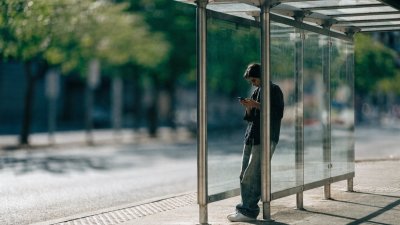 Pessoa encostada na estrutura de vidro de um abrigo de ônibus, usando o celular durante o dia